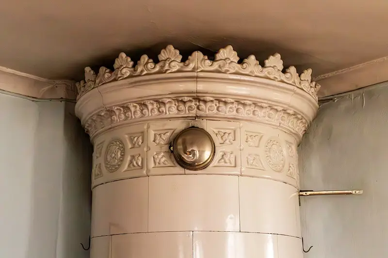 Detalj av en rund vit kakelugn med dekorerad krona och mässingslucka