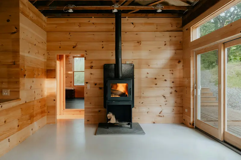 Liten svart vedkamin med eld bakom luckan på en stenplatta i ett litet rum med furupanel och glasdörrar