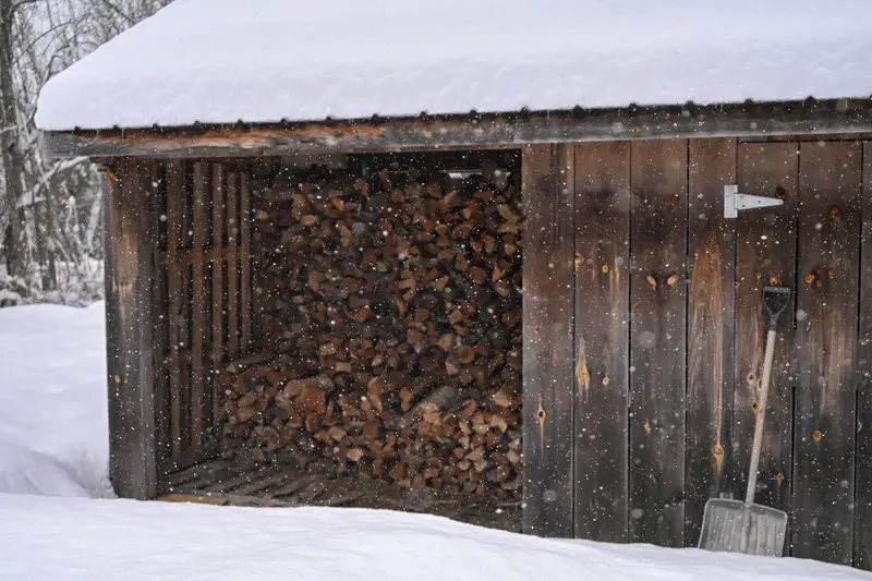 Vedbod av grått trä med snötäckt tak, fylld med staplad kluven ved, och en snöskyffel lutad mot väggen
